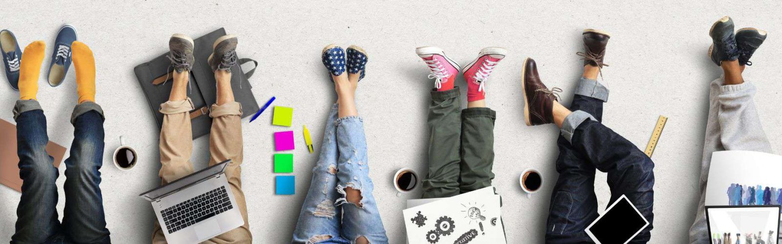Image of six people's legs sitting on the ground. With laptops, pens, rulers, coffee cups beside them all.