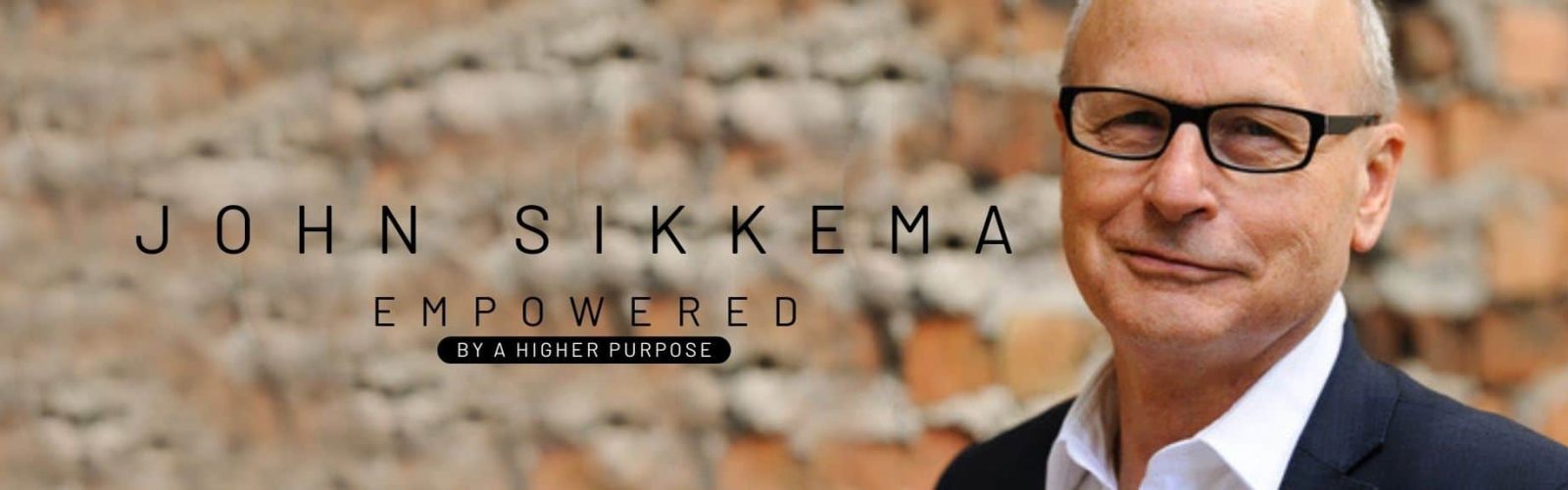 Image of a man in his fifties wearing a white shirt with black suit jacket and black framed glasses smiling. Behind his is a brick wall.