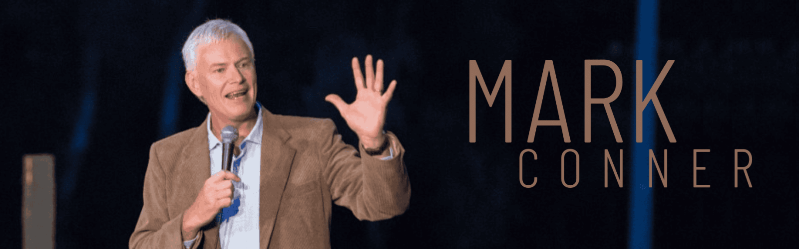 Mark Conner who is a man in his sixties is wearing a brown suit jacket and blue shirt holding a microphone presenting on stage.