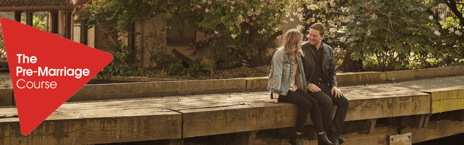 Image of a young man and women sitting by the water on a timbe walkway. They are looking at each other wearing jeans and casual jackets. The sun is shinning and their is a lovely garden behind them.